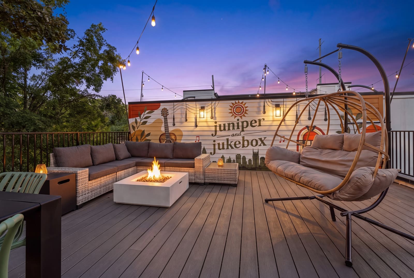 Rooftop deck at dusk with a fire pit, sectional and hanging egg chair in front of the Juniper and Jukebox mural
